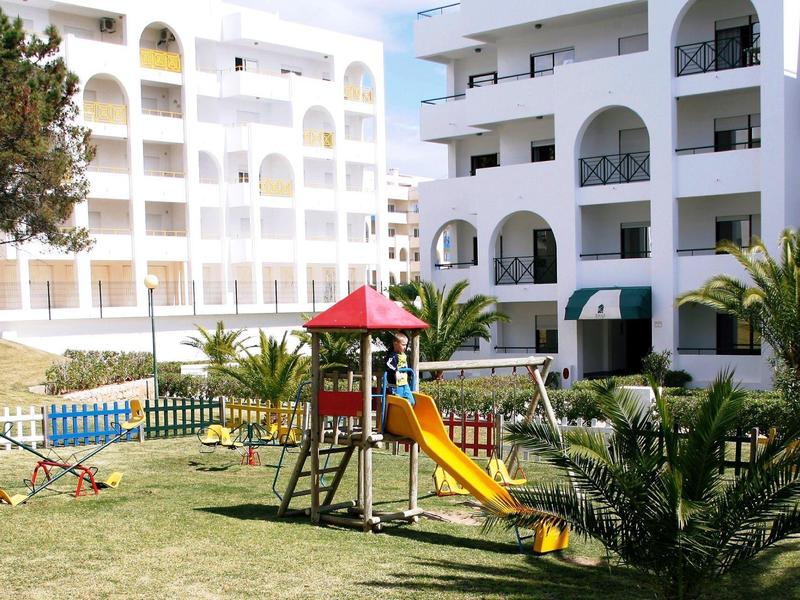 Spielplatz mit Rutsche vor weißen Wohn- oder Hotelgebäuden und grünem Rasen.