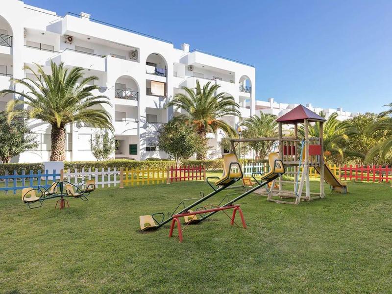 Grüner Spielplatz mit Schaukeln und Rutsche vor weißem Hotelgebäude und blauem Himmel.