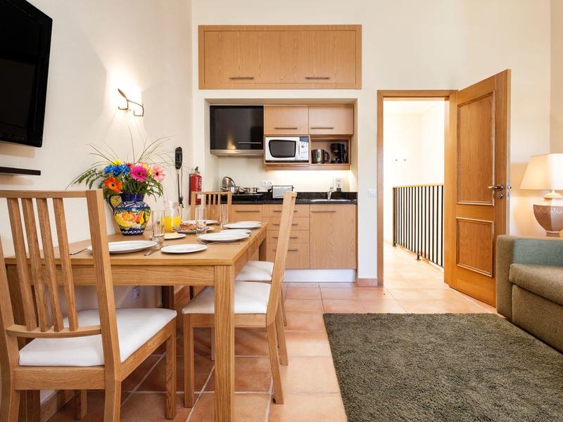 Gemütliche Wohnküche mit Holzmöbeln, Esstisch, grünen Teppich und Sofaecke in warmem Licht.