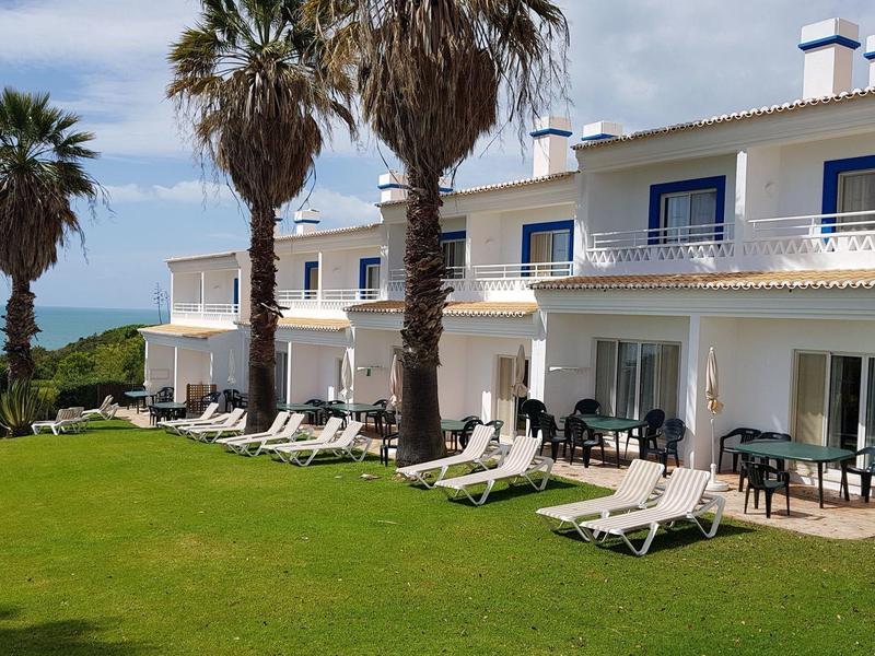 Weißes Haus mit blauen Fenstern, Liegestühlen und Palmen vor grünem Rasen am Meer.