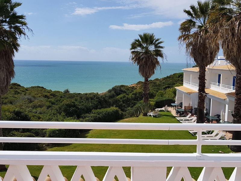 Blick von Balkon mit weißem Geländer auf Meer, Palmen und Gebäude bei klarem Himmel.