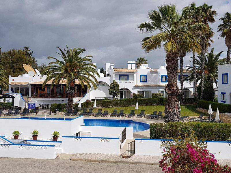 Weißes Hotel mit blauen Fenstern, Palme und blauem Pool unter bewölktem Himmel.