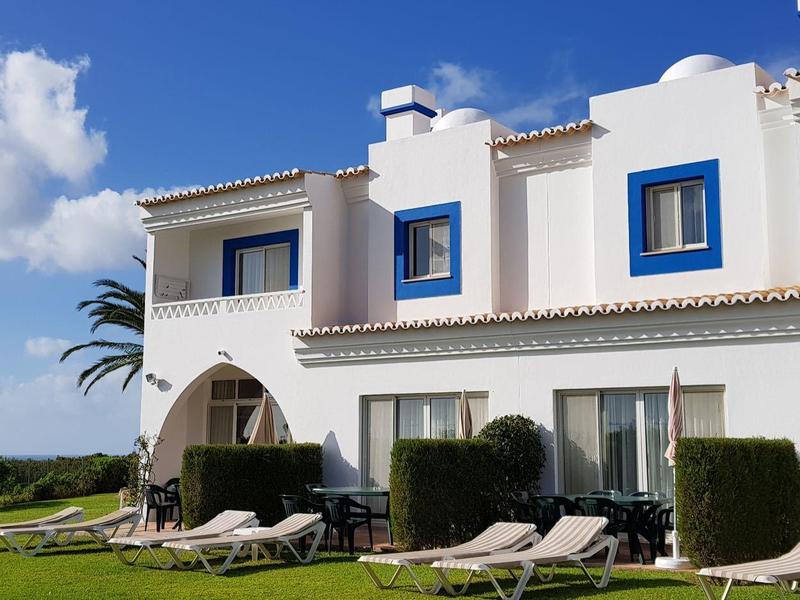 Ein großes weißes Haus mit blauen Fensterrahmen, grünen Büschen und Liegestühlen im Garten.