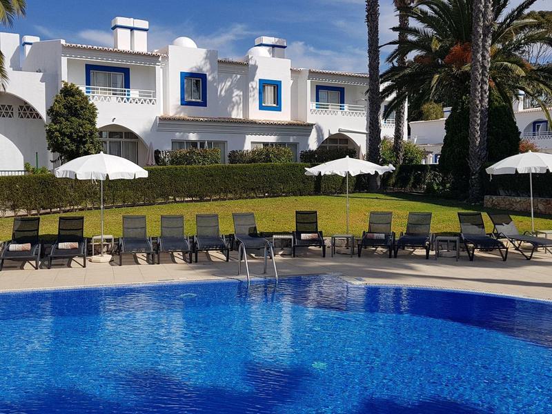 Großer blauer Pool vor weißen Häusern mit Palmen und Liegestühlen an einem sonnigen Tag.