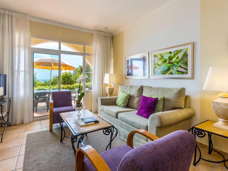 Helles Wohnzimmer mit Sofa, Sessel, Couchtisch, Bilderrahmen und Blick auf Terrasse mit Sonnenschirm.