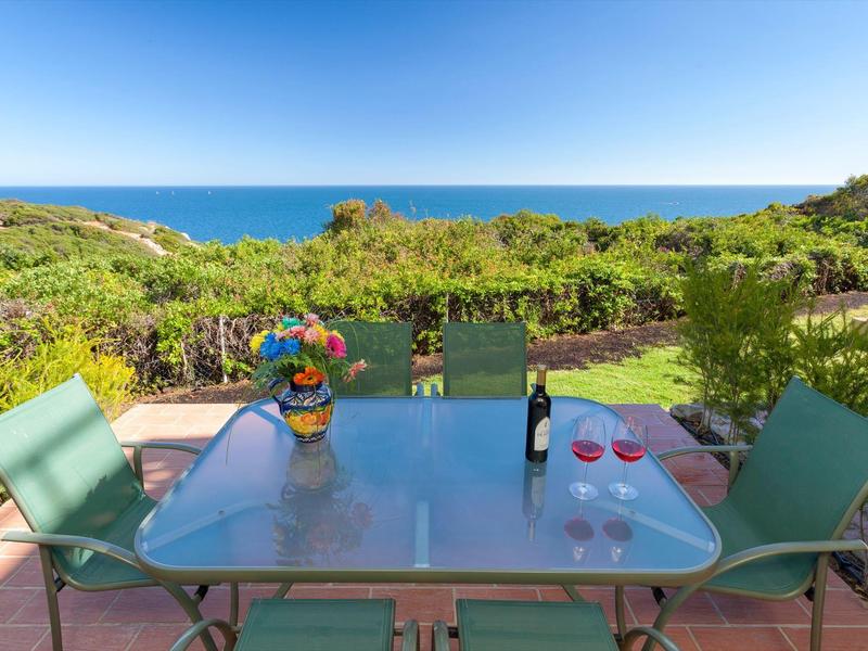 Terrasse mit blauem Glas-Tisch, grünen Stühlen, Blumenstrauß und Blick auf Meer und grüne Landschaft.