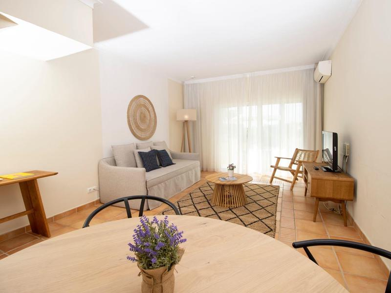 Helles Wohnzimmer mit grauem Sofa, Holzmöbeln, rundem Tisch und großen Fenstern mit Vorhängen.