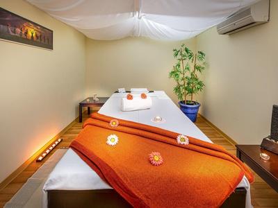 Gemütlicher Wellnessraum mit Massageliege, orangefarbener Decke, Kerzen und ruhiger Beleuchtung.