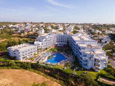 Großes Hotel mit weißer Fassade und blauem Pool, umgeben von Grün und Stadt im Hintergrund.