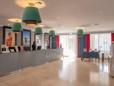 Moderne Hotellobby mit langer Rezeption, großen hellgrünen Hängelampen und beigen Fliesenboden.
