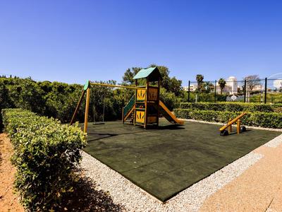 Kinderspielplatz mit Rutsche, Schaukel und Kletterhaus auf grünem Boden unter blauem Himmel.
