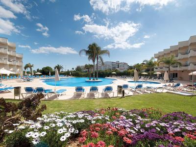 Großer Hotelpool mit blauen Liegestühlen, Palmen und bunten Blumen unter blauem Himmel.