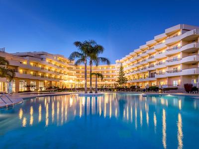 Ein beleuchtetes Hotel mit mehreren Stockwerken, Pool und Palmen bei Nacht.
