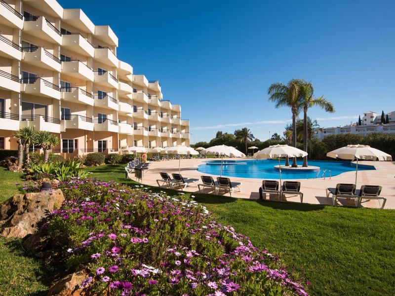 Moderner Hotelkomplex mit Swimmingpool, Liegestühlen, weißen Sonnenschirmen und blühenden Blumen.