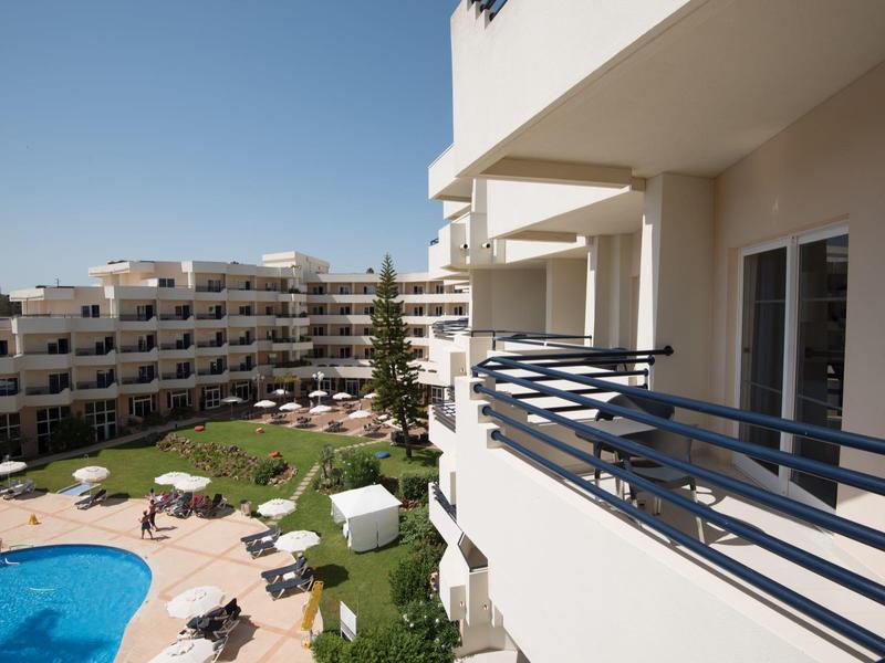 Blick von Balkon auf Hotelanlage mit Pool, Sonnenschirmen und mehrstöckigen Gebäuden bei klarem Himmel.