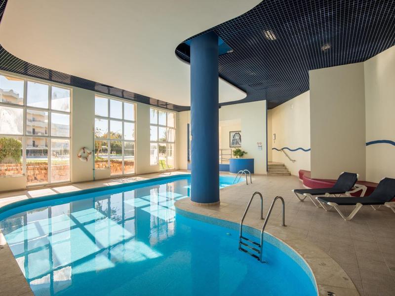 Innenbereich mit beleuchtetem Schwimmbecken, blauen Säulen, großen Fenstern und Liegestühlen.