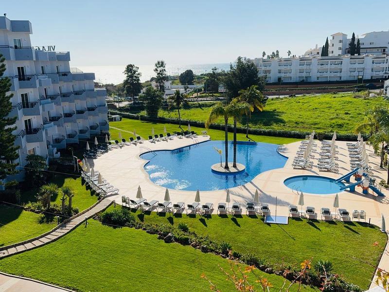 Großer Pool mit Sonnenschirmen, umgeben von grünen Rasenflächen und weißen Hotelgebäuden.