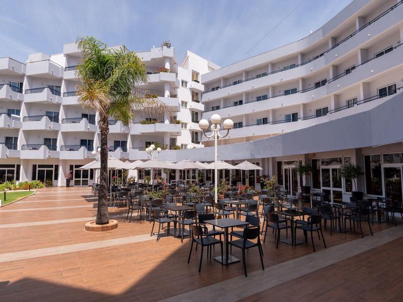 Großes, modernes Hotel mit weißer Fassade, Balkonzimmern, Sonnenschirmen und Außenbestuhlung.