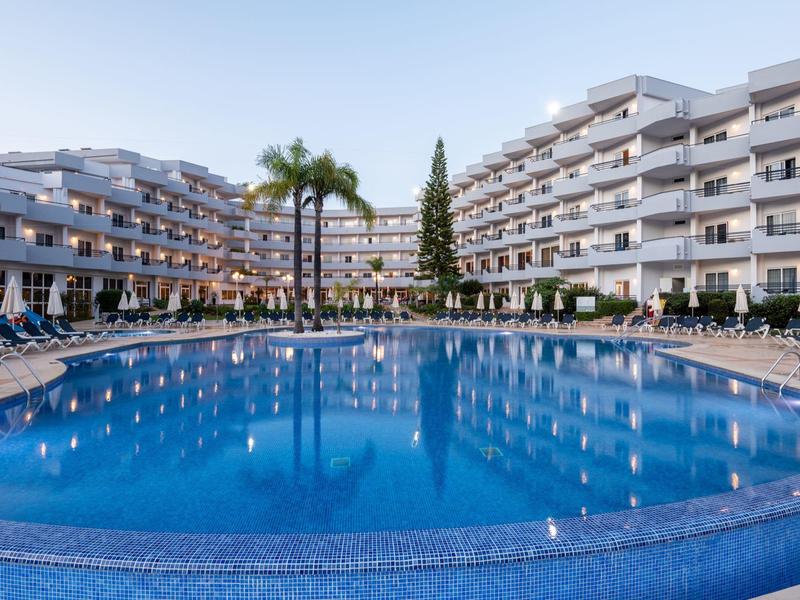 Großer blauer Pool mit vielen Liegestühlen vor modernen, weißen Hotelgebäuden und Palmen in Abendlicht.