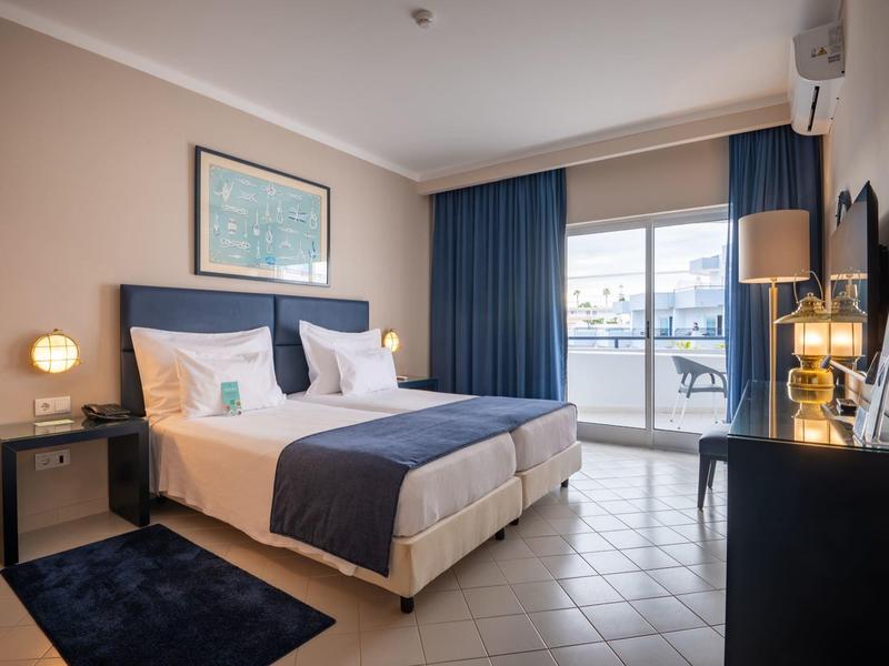 Helles Schlafzimmer mit Doppelbett, blauen Vorhängen und Zugang zu Balkon mit Blick nach außen.