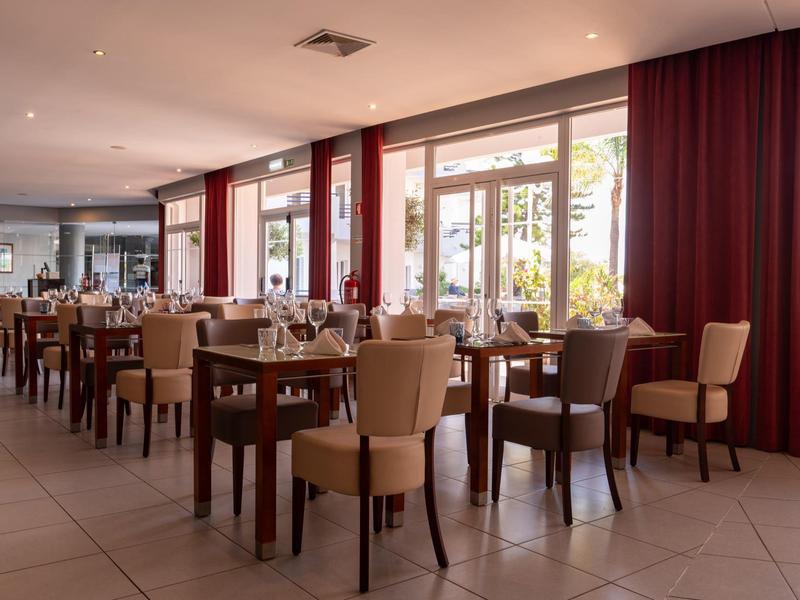 Ein heller, moderner Speisesaal mit vielen Holztischen und gepolsterten Stühlen neben großen Fenstern.