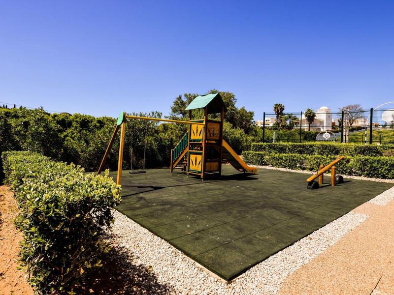 Kinderspielplatz mit Rutsche, Schaukel und Kletterhaus auf grünem Boden unter blauem Himmel.