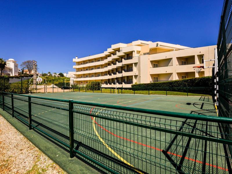 Tennisplatz mit grünem Belag vor mehrstöckigem gelbem Wohngebäude unter blauem Himmel.