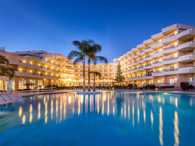 Großes Hotel mit beleuchtetem Pool, mehreren Stockwerken und Palmen bei klar blauem Himmel.