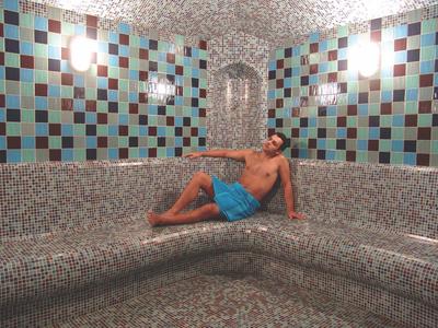 Ein Mann in blauer Badehose liegt entspannt auf einer grauen Mosaikbank in einem Raum mit bunten Kacheln.