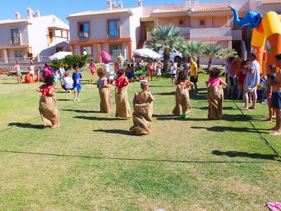 Kinder hüpfen in Säcken auf einer grünen Wiese vor bunten Häusern und einer Hüpfburg.