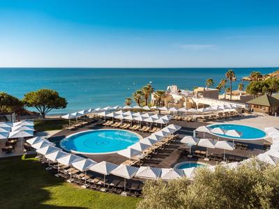 Poolanlage mit zwei runden Pools, weiß-blauen Sonnenschirmen und Blick aufs blaue Meer unter klarem Himmel.
