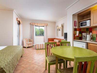 Helles Zimmer mit grünem Esstisch, Stühlen, Einzelschrankbett und bunter Küchenzeile.