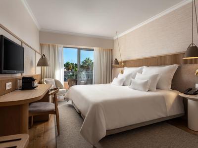 Modernes Hotelzimmer mit Doppelbett, Schreibtisch, Fernseher und Balkon mit Blick auf Palmen.