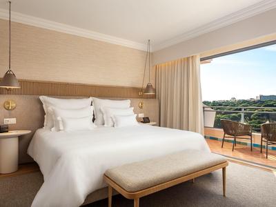 Helles Hotelzimmer mit Doppelbett, Holzmöbeln und Balkon mit Blick auf grüne Landschaft.