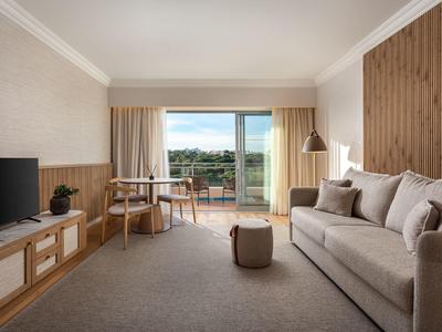 Helles Wohnzimmer mit grauem Sofa, Holzmöbeln, Teppichboden und Balkon mit grüner Aussicht.