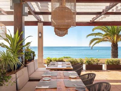 Terrasse mit Holztischen, Stühlen, grünen Pflanzen und Blick auf das blaue Meer und Palmen.