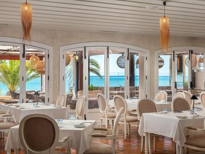 Helles Restaurant mit weißen Tischtüchern, eleganten Stühlen und Meerblick durch große Fenster.