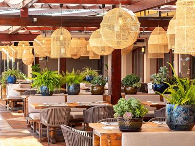 Offener Gastronomiebereich mit Holzmöbeln, grünen Pflanzen und warmen Lampenschirmen aus Weide.