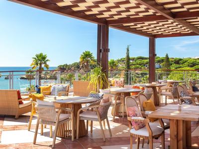 Terrasse mit Holzdach, Tischen und Stühlen, Blick aufs Meer und grüne Küste bei Sonnenschein.