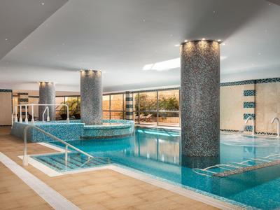 Hallenbad mit blauem Wasser, runden Säulen, Treppe und großen Fenstern, helle Decke und Wände.