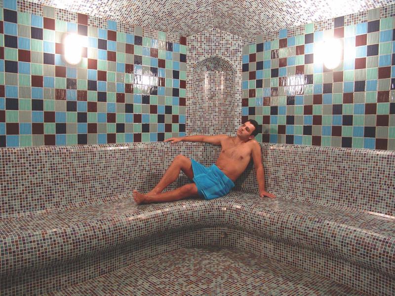 Ein Mann in blauer Badehose liegt entspannt auf einer grauen Mosaikbank in einem Raum mit bunten Kacheln.