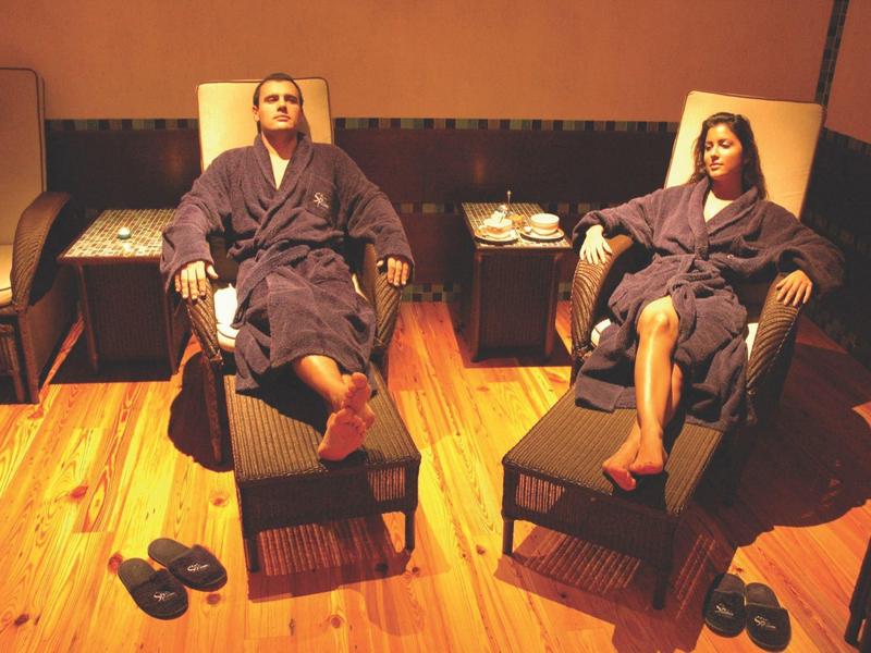 Ein Mann und eine Frau in Bademänteln entspannen auf Liegestühlen in einem warm beleuchteten Raum.