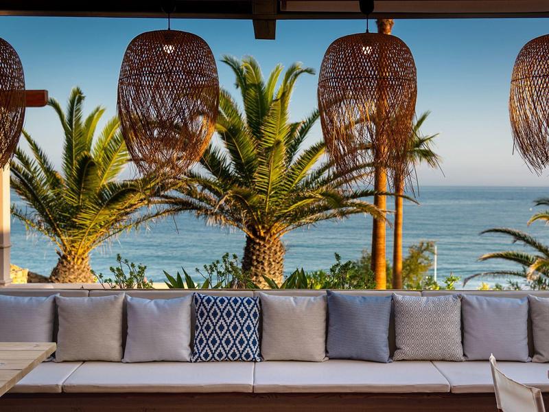 Gemütliche Outdoor-Lounge mit grauen Kissen, Palmen und Meerblick unter geflochtenen Lampen.