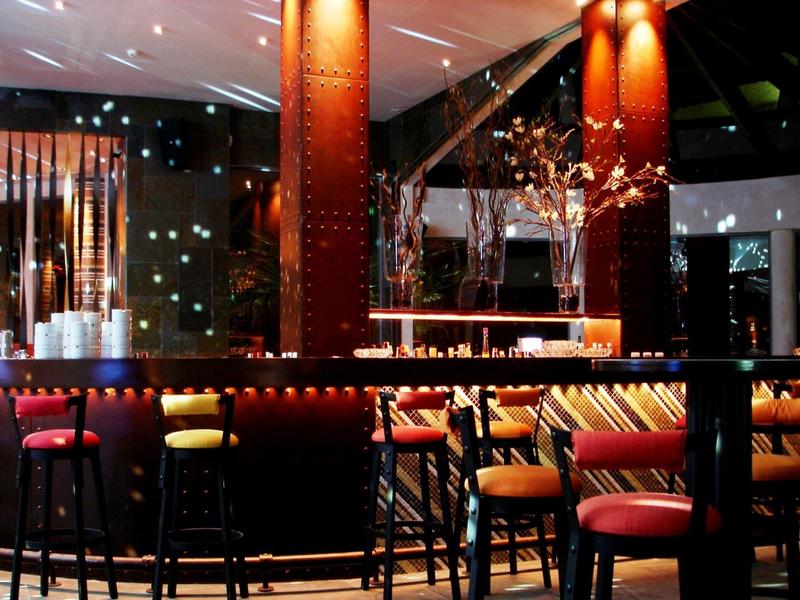 Elegante Bar mit hohen Stühlen, warmem Licht und Kerzen auf dunklen Tischen in modernem Ambiente.