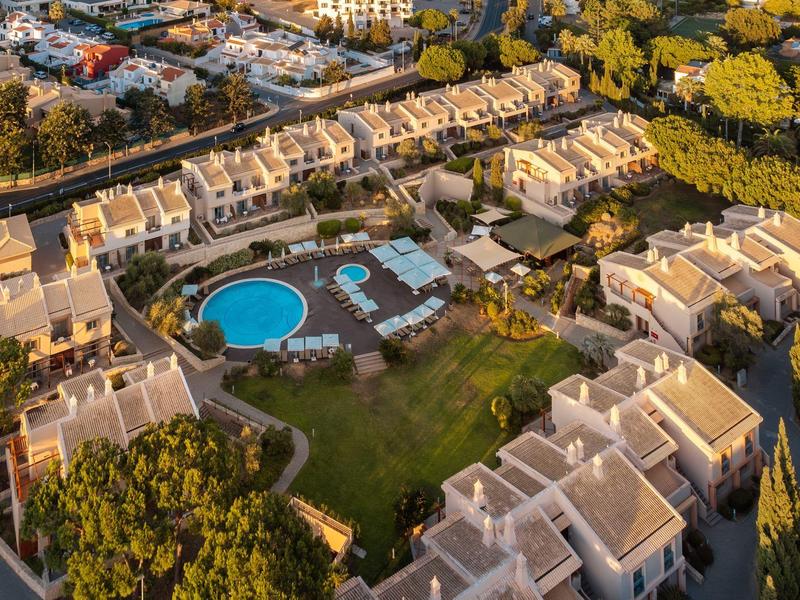 Wohnanlage mit mehreren weißen Gebäuden, rundem blauen Pool und grünen Bäumen bei Sonnenaufgang.