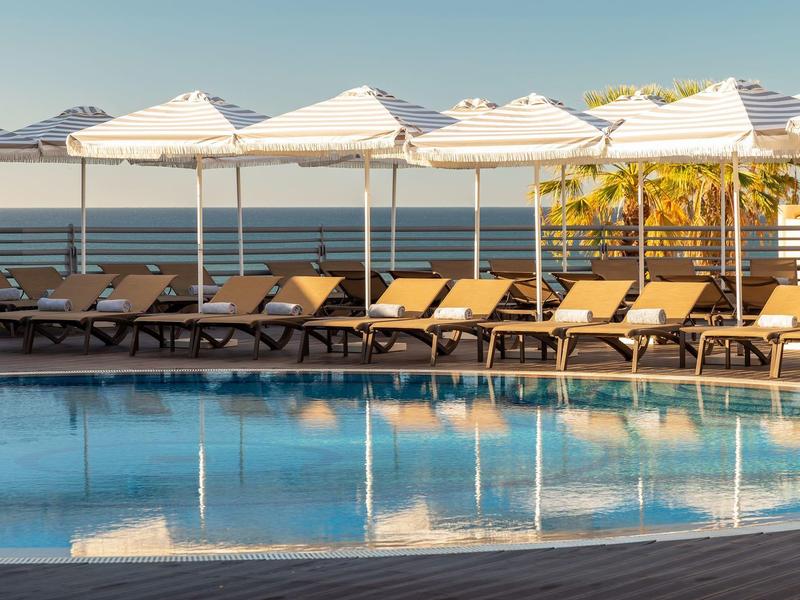 Pool mit vielen Liegestühlen und weißen Sonnenschirmen am ruhigen Wasser unter blauem Himmel.