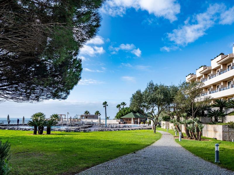 Blick auf gepflegten Garten, Weg, Palmen und Gebäude unter blauem Himmel mit Wolken.