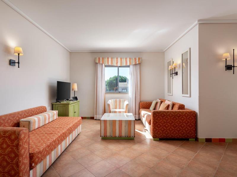 Hell eingerichtetes Wohnzimmer mit orangen Sofas, kleinem Tisch, TV und Fenster mit Vorhang.