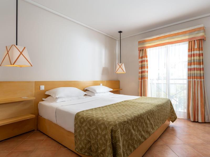 Helles Schlafzimmer mit zwei Einzelbetten, Holzmöbeln, beigem Bettüberwurf und karierten Vorhängen.