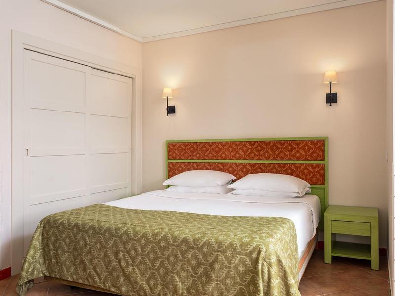 Schlafzimmer mit Doppelbett, grün gemustertem Bettüberwurf, rotem Kopfteil und grünem Nachttisch.